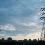 A city skyline at dusk with transmission towers and power lines silhouetted against a cloudy sky.