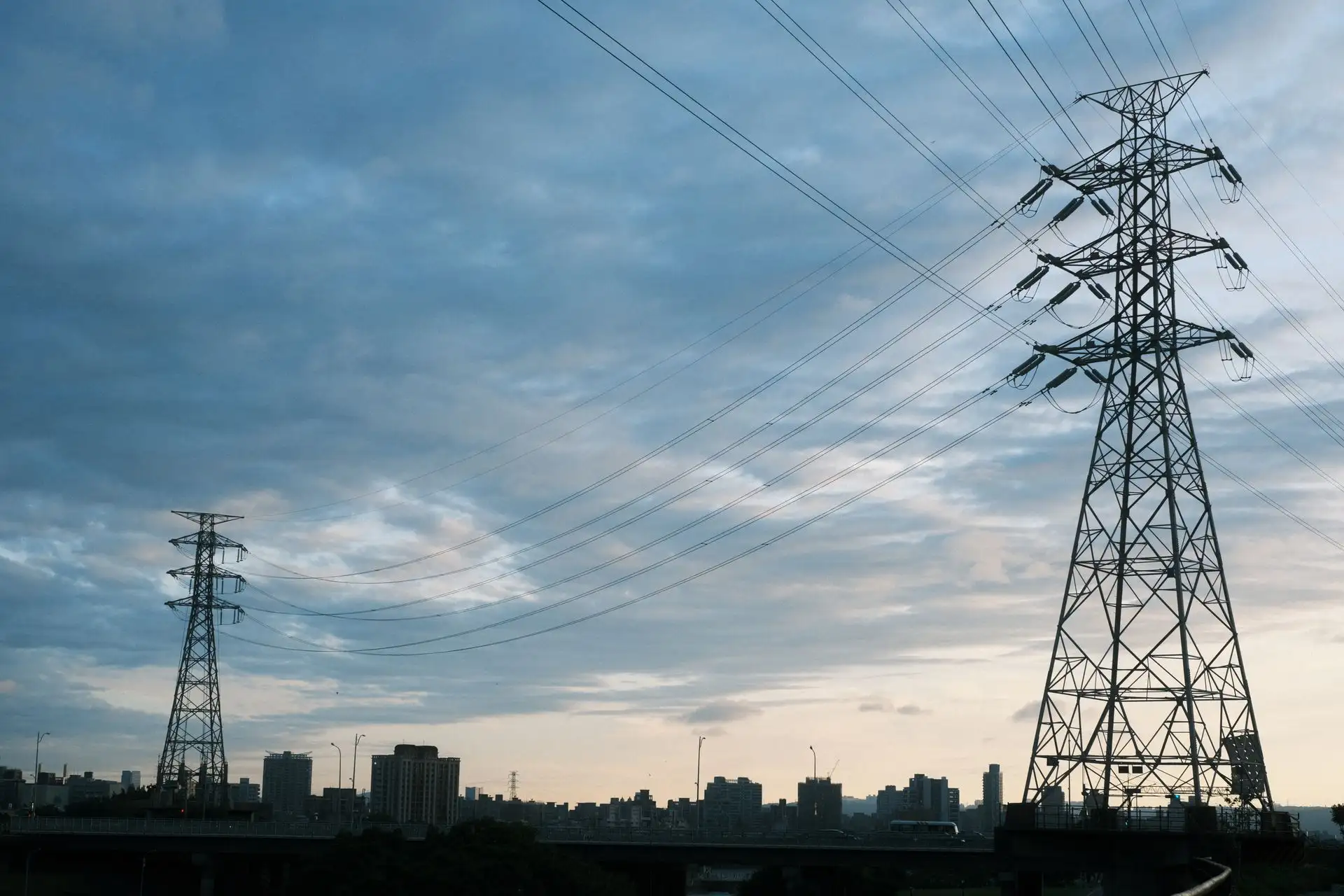 A city skyline at dusk with transmission towers and power lines silhouetted against a cloudy sky.
